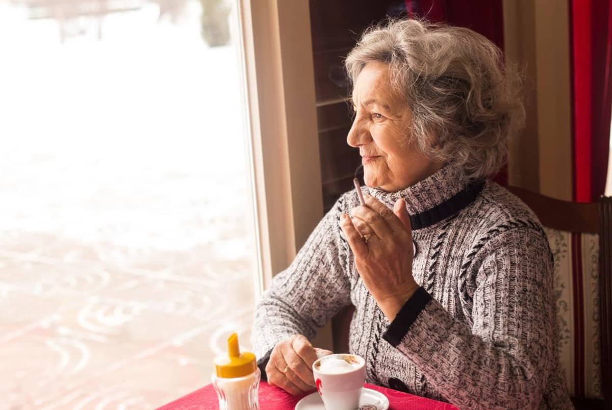 A senior woman is sitting smoking a cigarette and drinking coffee. She is looking out window.